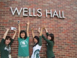 four girls in spartan apparel with their arms up in front of a red brick building named 'Wells Hall'